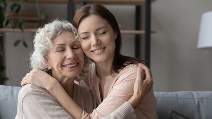 Embracing mommy. Affectionate elderly mother and grown up daughter sit on couch close together with closed eyes hug touch heads. Old mom and adult child hold each other in arms reconcile after quarrel