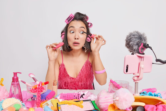 Young Asian lady applies mascara presents new beauty product records livestreaming video sits at messy table wears hair rollers pink dress isolated over white background. Professional female artist - Powered by Adobe
