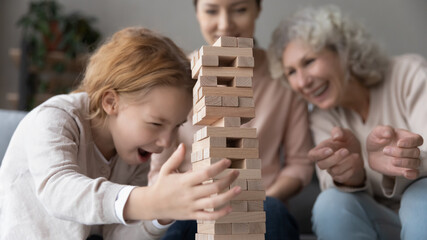 Cute game. Young adult mum and retired elderly granny play together with preteen daughter grandkid building brick construction on table. Focus on little girl trying to stop wooden tower from falling