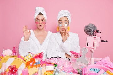 Surprised diverse women wear white bathrobe and towels over head give review creat video about skin care apply beauty patches pose near table with cosmetic products around. Blogging concept.