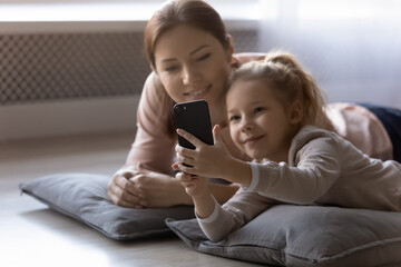 Floor heating. Relaxed adult elder sister take rest on pillows with younger one little girl watch cute video clip on cellphone together. Small adopted daughter show image on phone screen to foster mom