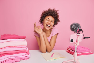 Obraz na płótnie Canvas Happy curly female blogger thinks about new content smiles pleasantly writes down information in notebook folds laundry uses smartphone webcam isolated over pink background. Monochrome shot.