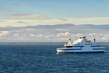 Fototapeta na wymiar The small car ferry runs between Virtsu harbour Estonia and Saaremaa Island on Baltic Sea. Calm sea and blue sky with white clouds. In the background the wind farm on the seashore.