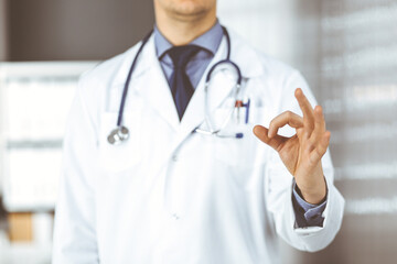 Unknown male doctor standing straight with OK sign in clinic near his working place, closeup. Perfect medical service in hospital. Medicine and healthcare concept