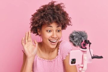 Indoor shot of cheerful woman with curly Afro hair waves hello in smartphone camera talks with subscribers records educational video poses in front of modern gadget on tripod against pink wall