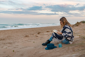 a girl with a blue hat is taking a coffee watching the sea while is reading a book