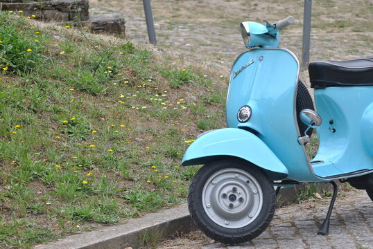Piaggio Vespa azul parada numa rua de uma aldeia antiga, lambreta, mota italiana, modelo cl&aacute;ssico dos anos 60, jardim com flores