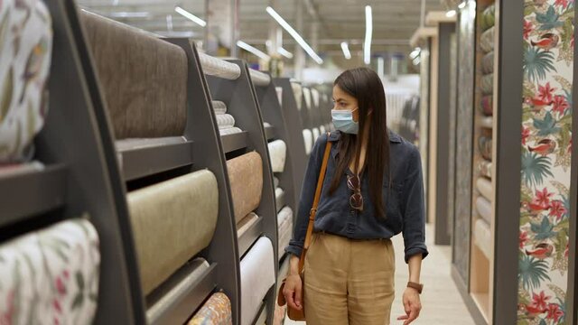 Young woman wearing face protective mask choosing wallpapers for home repair while doing shopping in construction department of big hypermarket during coronavirus epidemic