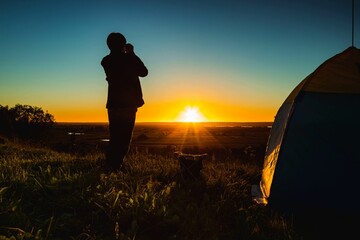 Silhouette of a man with binoculars near the tent against the backdrop of the sunset on a clear sky over the meadow.