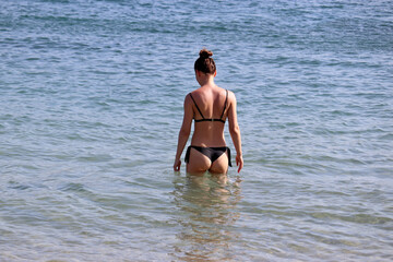Slim girl in bikini going to swim in sea water, rear view. Beach vacation, summer leisure