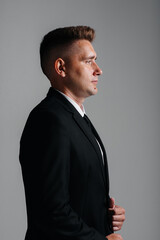 A stylish young man in a business suit poses sideways on a gray background. A handsome charismatic businessman.