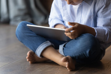 Gen Z kid sitting on heating floor at home, using learning app on tablet, doing school homework, watching video, reading book, browsing internet. Smart primary school kid studying gadget