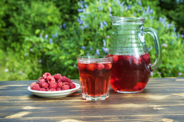 A jug and a mug with compote and a plate with fresh raspberries on a wooden table.