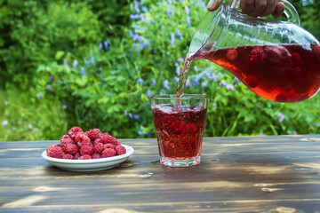 The compote is poured from a glass jug into a mug. A plate of ripe raspberries on a wooden table
