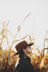 Beautiful stylish woman in brown hat and vintage coat relaxing in autumn maize field in sunset light. Portrait of fashionable young hipster female in retro outfit enjoying evening in autumn corn