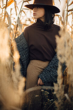Beautiful stylish woman in brown hat and vintage coat posing in autumn maize field in warm sunny light. Fashionable young hipster female standing in autumnal corn in evening countryside