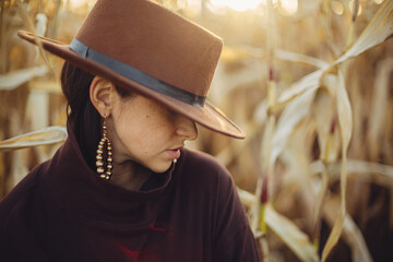 Portrait of stylish woman in hat and brown clothes posing in autumn maize field in warm sunny...