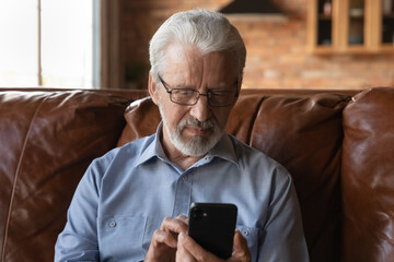 Focused elder 70s man using telemedicine app on smartphone at home, looking at screen, reading text, browsing internet, making video call for distance talk to doctor, shopping online. - Powered by Adobe