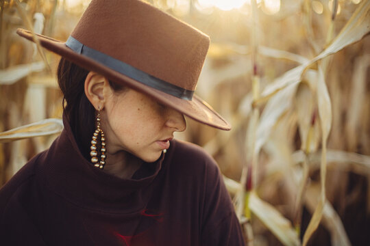 Portrait of stylish woman in hat and brown clothes posing in autumn maize field in warm sunny light. Fashionable attractive young female  standing in autumnal corn in evening countryside