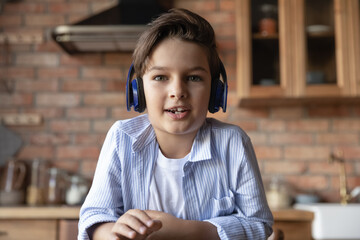 Portrait of cute gen Z school boy in headphones speaking at webcam during video call. Kid learning online, studying from home, using gadget for virtual talk to teacher. Screen view head shot