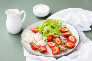 Fresh whole and sliced strawberries, cottage cheese and lettuce on a plate and sour cream in a bowl on a green background. Summer vitamin snack