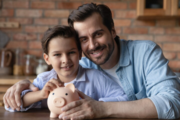 Happy dad and little son holding piggy bank, smiling, looking at camera. Father and kid saving...