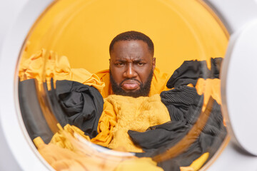 Discontent dark skinned bearded male householder does washing poses through washing machine drum does housework has unhappy face expression as doesnt want to do laundering. Laundry time concept