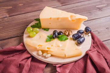 Fresh tasty sliced hard cheese garnished with herbs on a round wooden board on a dark wooden background