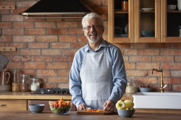 Happy senior 70s man wearing apron, cooking dinner in stylish kitchen, cutting fresh organic...