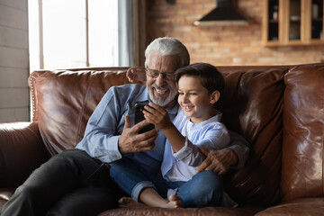 Happy preschooler boy and mature senior 70s grandpa using smartphone together, making video call, taking selfie, smiling at screen, enjoying leisure together with gadget at home, talking to family