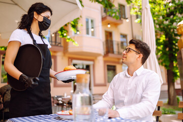 Young waitress in a protective medical mask and gloves with ordered meals serves food at table outside. Coronavirus outbreak lifestyle. Social distancing.