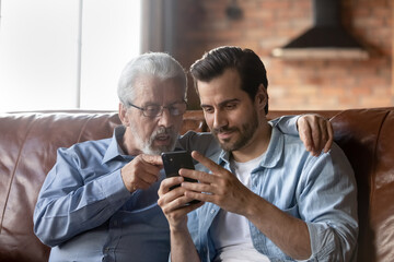 Grown son teaching elderly 70s dad to use mobile app on smartphone, showing family pictures,...