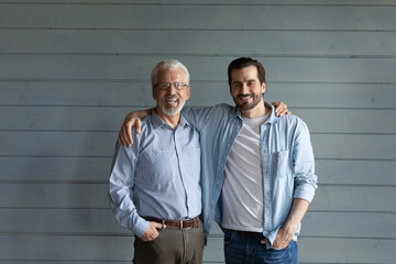 Happy mature senior 70s father and grown son standing together, hugging, looking at camera with...