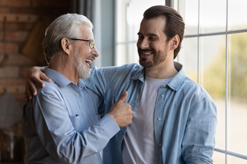 Fototapeta na wymiar Happy proud mature older father expressing approval, showing thumb up like gesture to excited grown son. Two family generation men meeting at home, talking, hugging, laughing together