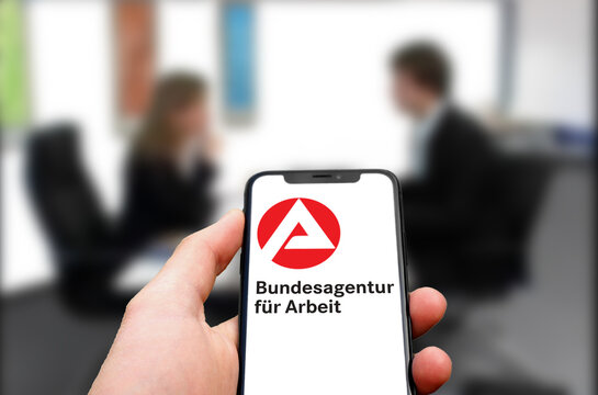Bundesagentur f&uuml;r Arbeit Logo wird von einem modernen Smartphone angezeigt, im Hintergrund ein Vorstellungsgespr&auml;ch im B&uuml;ro, Stellenb&ouml;rse, Karriere, Bewerbungsgespr&auml;ch, 2021