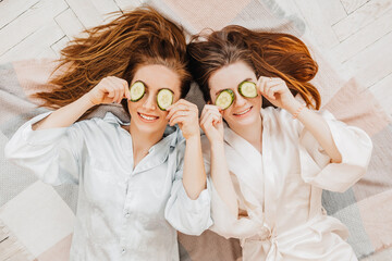 Two girls make homemade face and hair beauty masks. Cucumbers for the freshness of the skin around the eyes. Women take care of youthful skin. Girlfriends laugh at home lying on the floor on pillows.