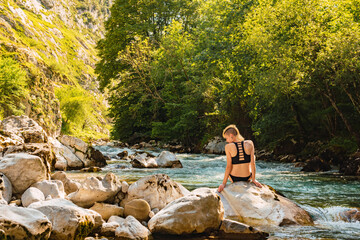 Fototapeta na wymiar Young Caucasian woman in bikini sitting on a rock near the river. Landscape with a river going through a forest. National Park of Picos de Europa and Rio Cares.