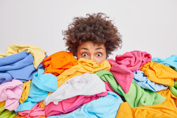 Shocked woman with curly Afro hair stares bugged eyes drowned in huge pile of colorful clothing...