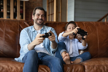Happy excited preschooler kid and dad playing video game at home, using joysticks, sitting on...