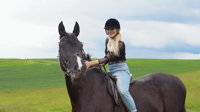 Glamorous young woman in a helmet, trendy blouse with translucent lush sleeves and light jeans sits on a black horse in the field. The rider strokes the horse's neck