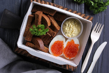 Healthy food for delivery. Breakfast. Boiled eggs with red caviar, crackers and yellow sauce and herbs in a white lunch box on a wooden board on a dark gray background. Rustic. Background image