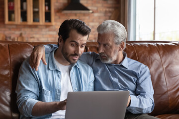 Fototapeta premium Grown son teaching senior father to use online apps and payment services on laptop. Two family generations men with computer sitting on couch together, browsing internet, talking, discussing content
