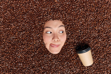 Young Asian woman coffee lover looks at appetizing cup of refreshing beverage licks lips with tongue surrounded by brown roasted seeds containing high amput of antioxidants. Source of caffeine