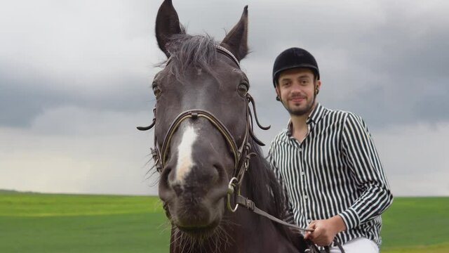 A young man in a helmet, striped black and white shirt and light trousers sits on a black horse in the field. Equestrian therapy. Horseback riding lessons. A walk on a horse