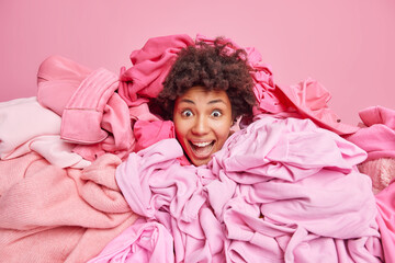 Overjoyed curly haired woman covered with pile of laundry sotrs out clothes in closet buys clothing on big sales being addicted shopaholic looks happily at camera isolated over pink background