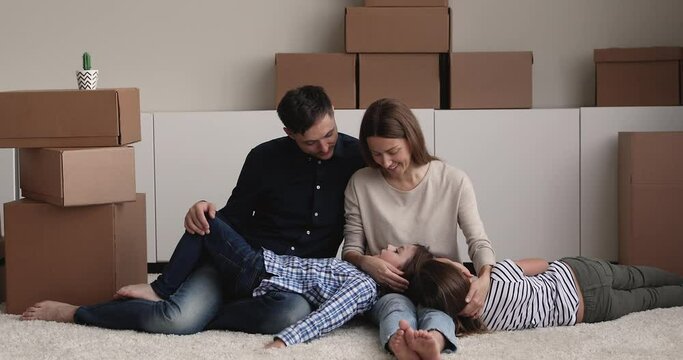 Young parents their kids rest on carpet in unfurnished living room with heap of carton boxes at relocation day, loving family talking enjoy repose at new own house. Bank loan, future, mortgage concept