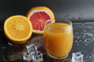 The juice of orange abicos yellow in the glass next to lie half of orange and grapefruit ice on a black background. Refreshing Drinks Summer