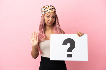 Young mixed race woman with pink hair isolated on pink background holding a placard with question mark symbol and doing stop sign
