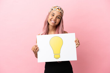 Young mixed race woman with pink hair isolated on pink background holding a placard with bulb icon with happy expression