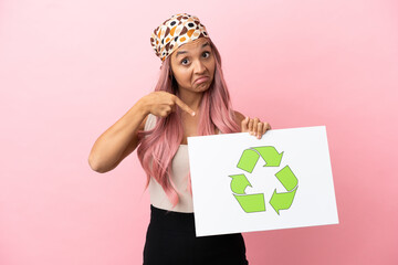 Young mixed race woman with pink hair isolated on pink background holding a placard with recycle icon and  pointing it